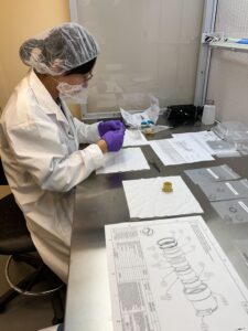 Dr. Shaojie Chen sits at a steel desk in a lab coat, mask, and hair net, with a lens in her hands. There are diagrams of the instrument on the desk in front of her.